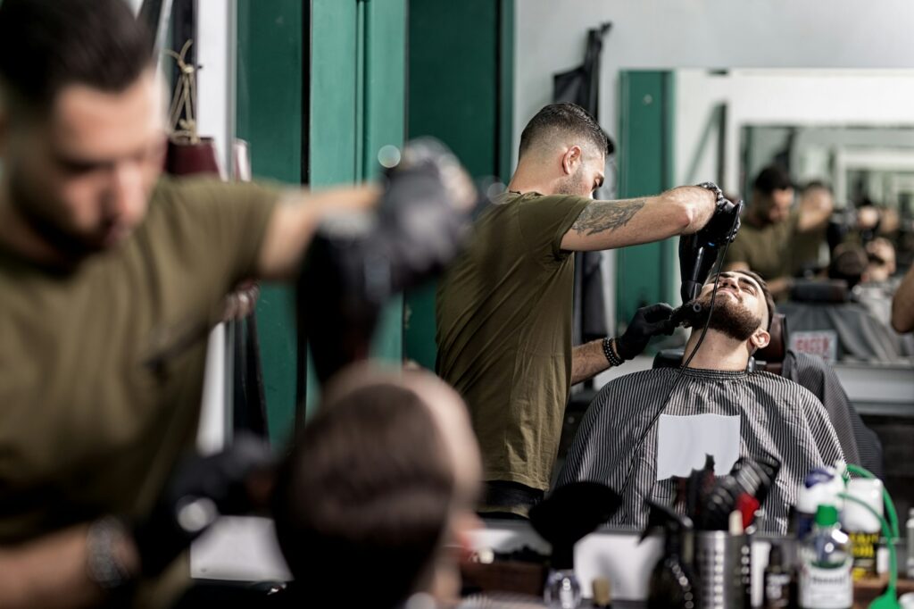 Brutal man with a beard sits in front of the mirror at a barbershop. Barber trims mens beard with